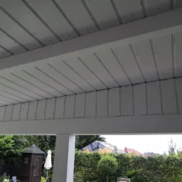 Biała, drewniana konstrukcja zadaszenia tarasu, widok od dołu, z widocznymi elementami mocującymi i fragmentem ogrodu w tle.