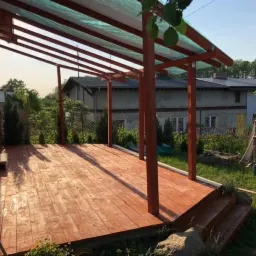 Drewniany taras z zadaszeniem w trakcie budowy, widoczne krokwie i deski podłogowe, w tle dom i zieleń.