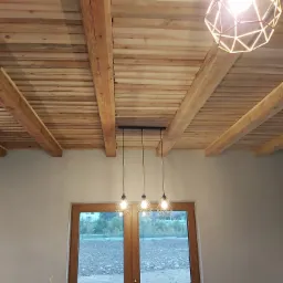 Drewniany sufit z belkami i trzema lampami wiszącymi nad oknem z widokiem na zewnątrz, dodatkowo jedna lampa w geometrycznej obudowie w prawym górnym rogu.