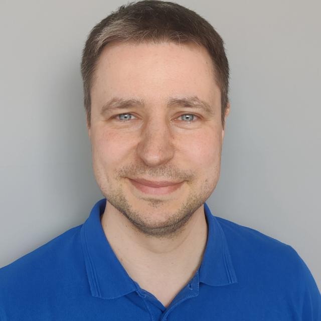 Portret mężczyzny o niebieskich oczach, ubranego w niebieską koszulkę polo, na jednolitym szarym tle. Widoczny delikatny uśmiech i krótko ostrzyżone włosy.