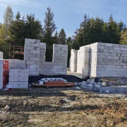 Wznoszone ściany domu z bloczków Termobet na fundamencie, widoczny stos zapakowanych bloczków, rusztowanie i drzewa w tle.