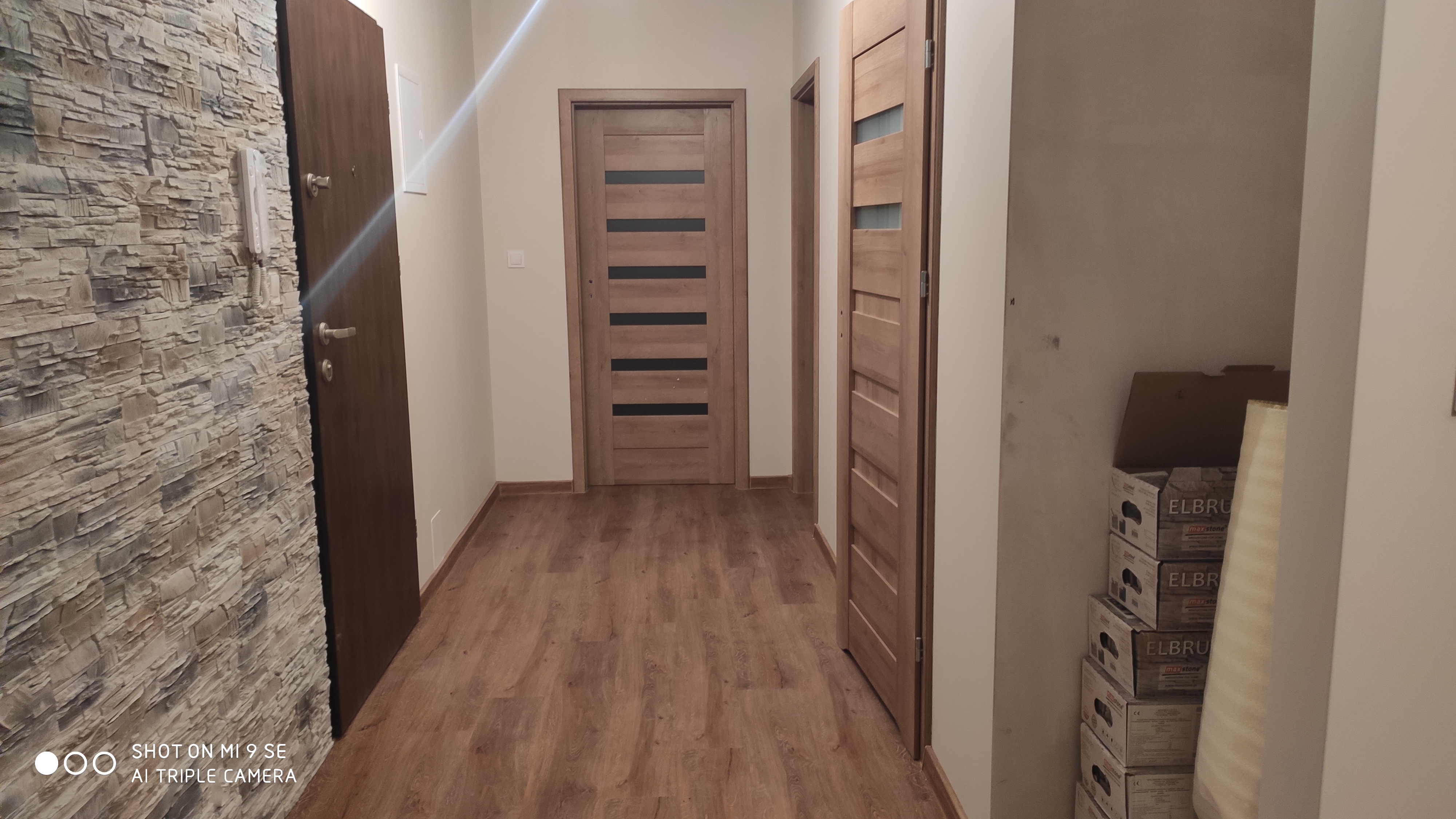 Korytarz po remoncie z panelami podłogowymi imitującymi drewno, okładziną ściany z kamienia dekoracyjnego, trzema parami drzwi wewnętrznych i stosem kartonów z płytkami pod ścianą.