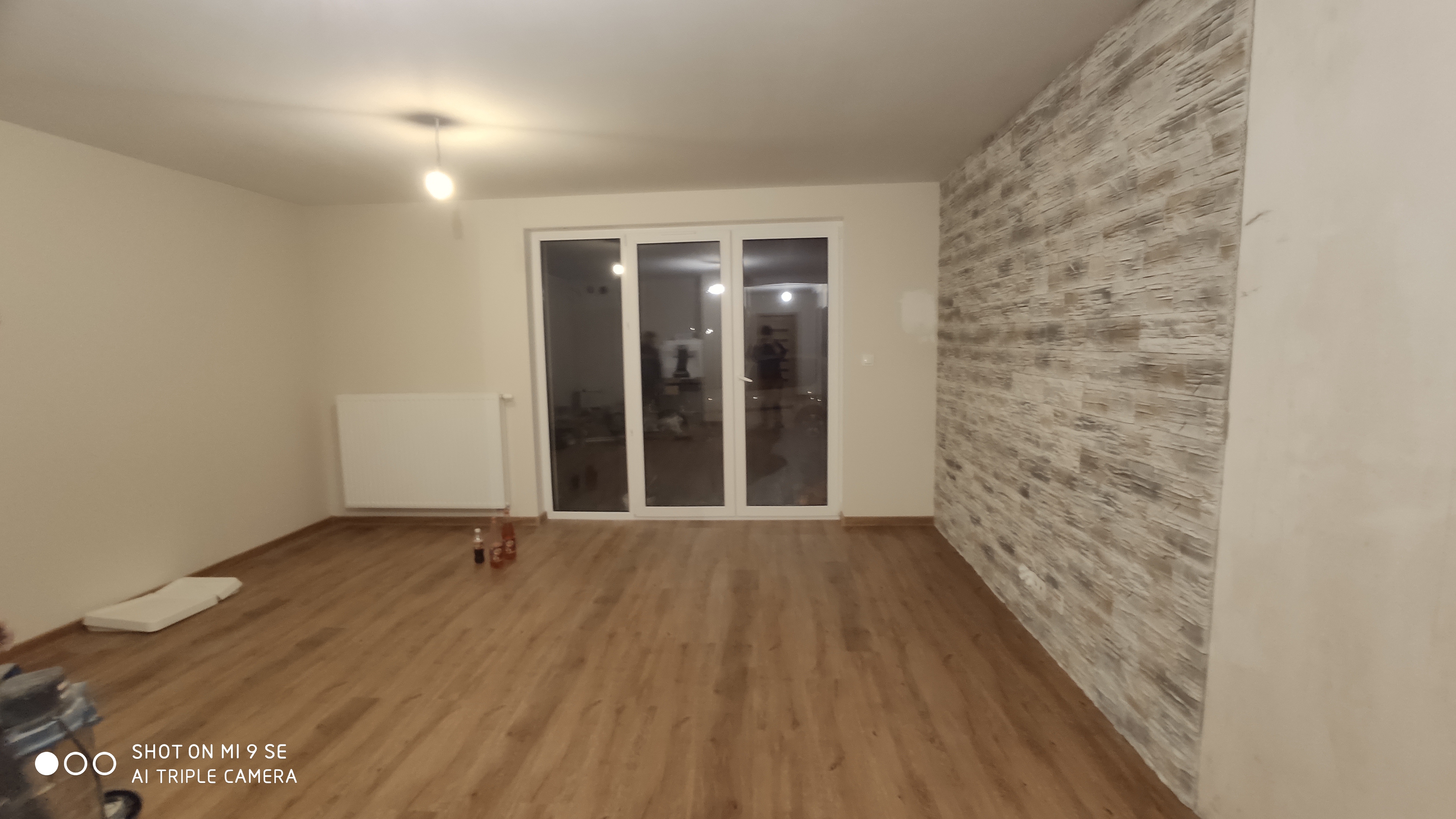 Jasne wnętrze pokoju z drewnianą podłogą, ścianą wyłożoną imitacją kamienia i dużymi przesuwnymi drzwiami balkonowymi, widok na nocne oświetlone osiedle.