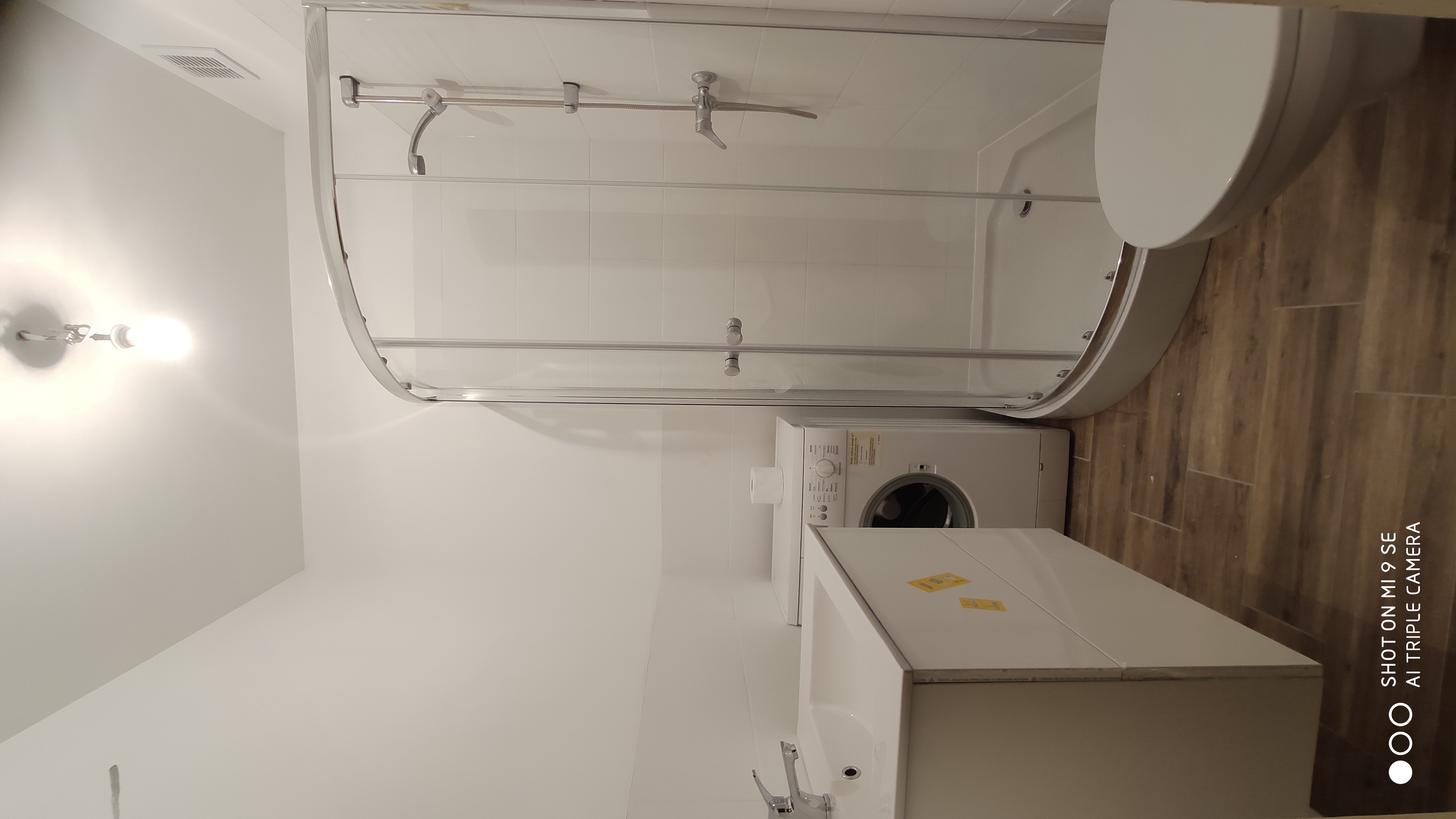 Biała łazienka z kabiną prysznicową z zaokrąglonymi drzwiami, umywalką z szafką, pralką i podłogą imitującą drewno.