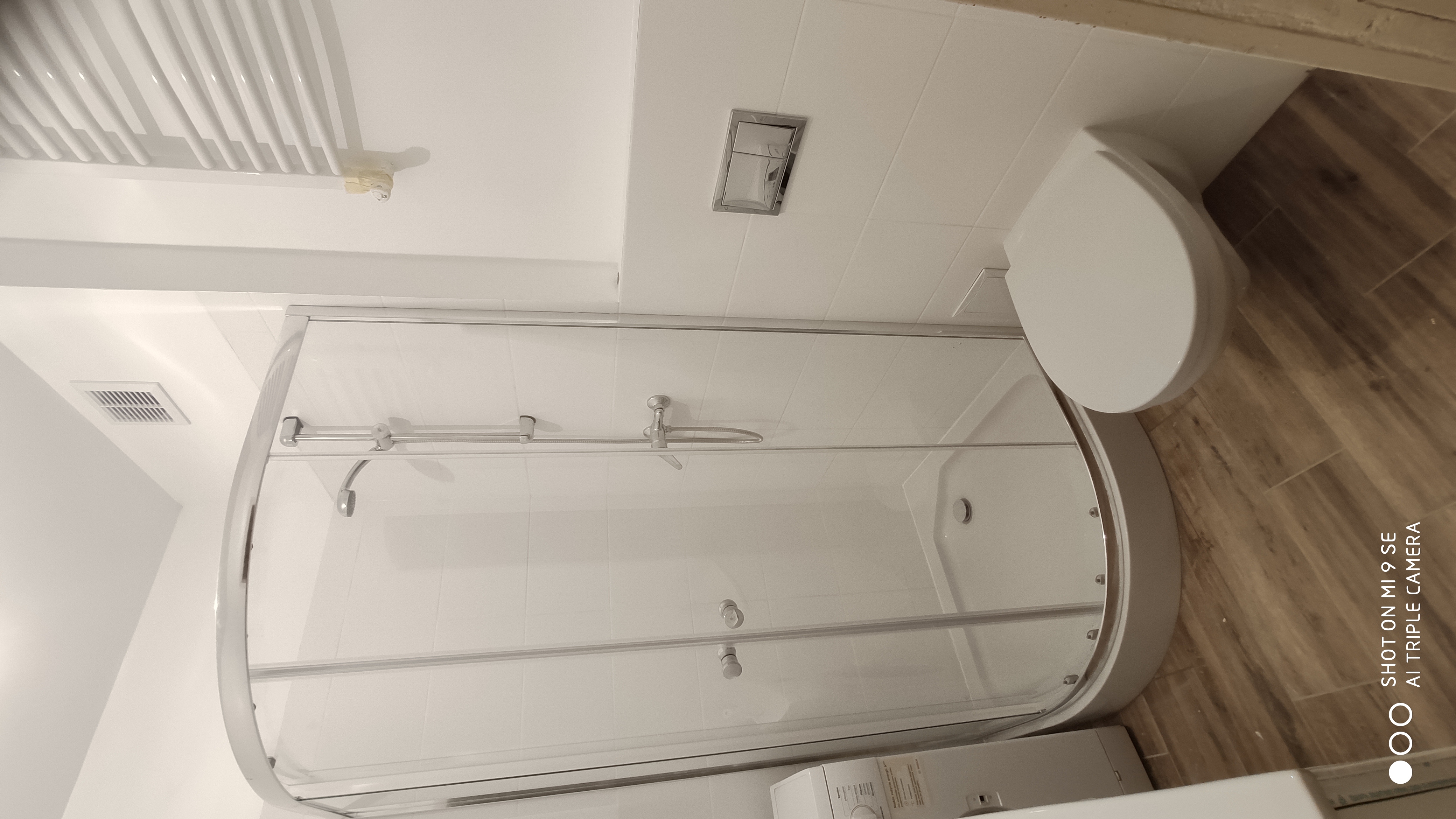 Nowoczesna łazienka z kabiną prysznicową z zaokrąglonym szkłem, podwieszaną toaletą z chromowanym przyciskiem spłukiwania i grzejnikiem drabinkowym na białej ścianie z płytek, uzupełniona podłogą...