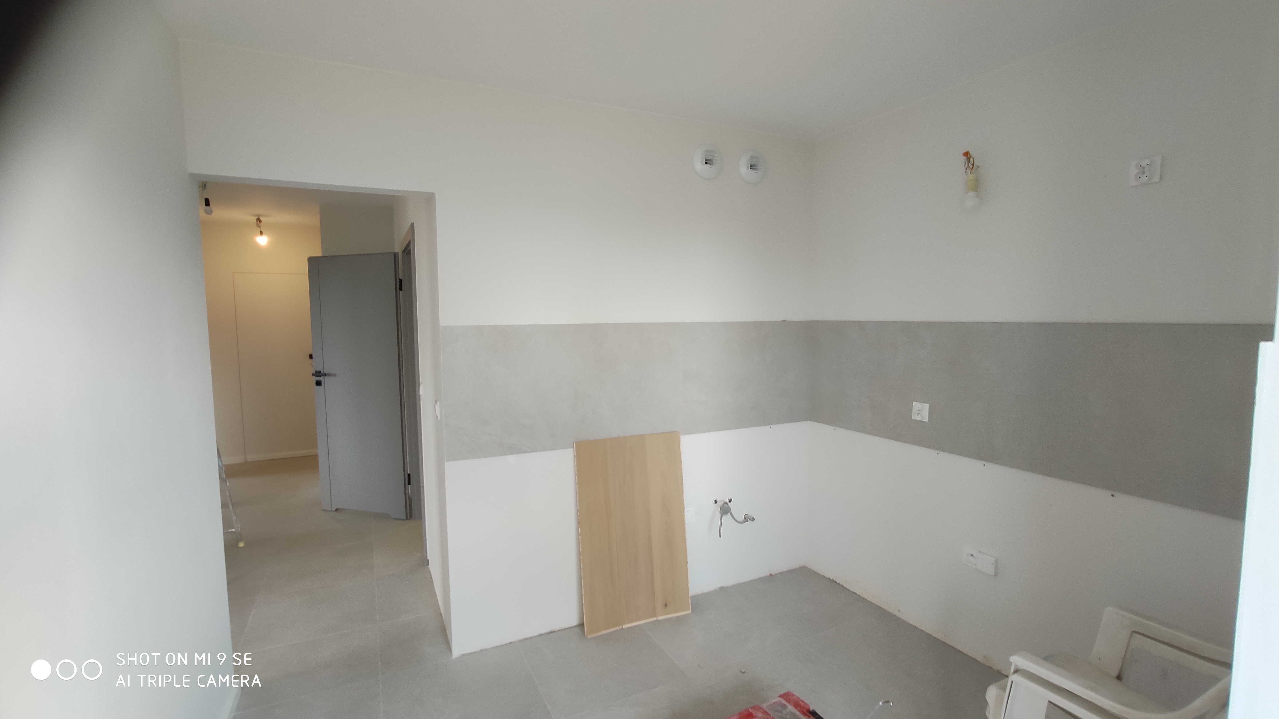 Wnętrze pomieszczenia w trakcie prac wykończeniowych, widoczne szare płytki częściowo ułożone na ścianach, odsłonięte instalacje wodne i elektryczne, oraz płyta z jasnego drewna oparta o ścianę...