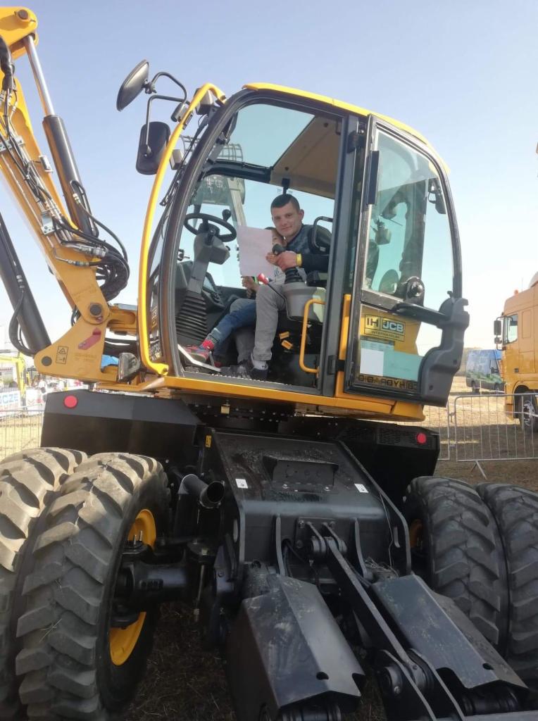 Żółto-czarna koparka kołowa JCB z operatorem i dzieckiem w kabinie, trzymającymi kartkę. Widoczne opony, ramię koparki i kabina z logiem JCB. Tło rozmyte, ekspozycja zewnętrzna.