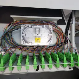 Rozdzielnica światłowodowa z licznymi kolorowymi kablami zakończonymi zielonymi złączkami, widoczny znak ostrzegawczy lasera na białej kasecie spawów.