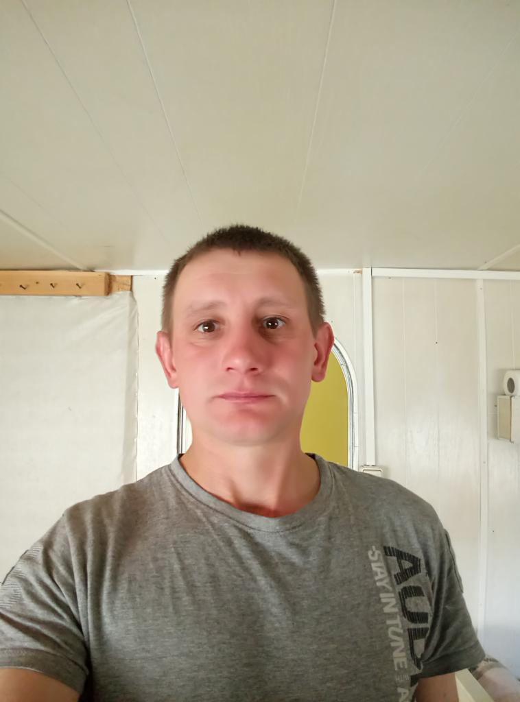 Mężczyzna w szarej koszulce na tle wnętrza z białą boazerią i widocznym fragmentem drzwi. Widoczne listwy przypodłogowe i fragment sufitu.