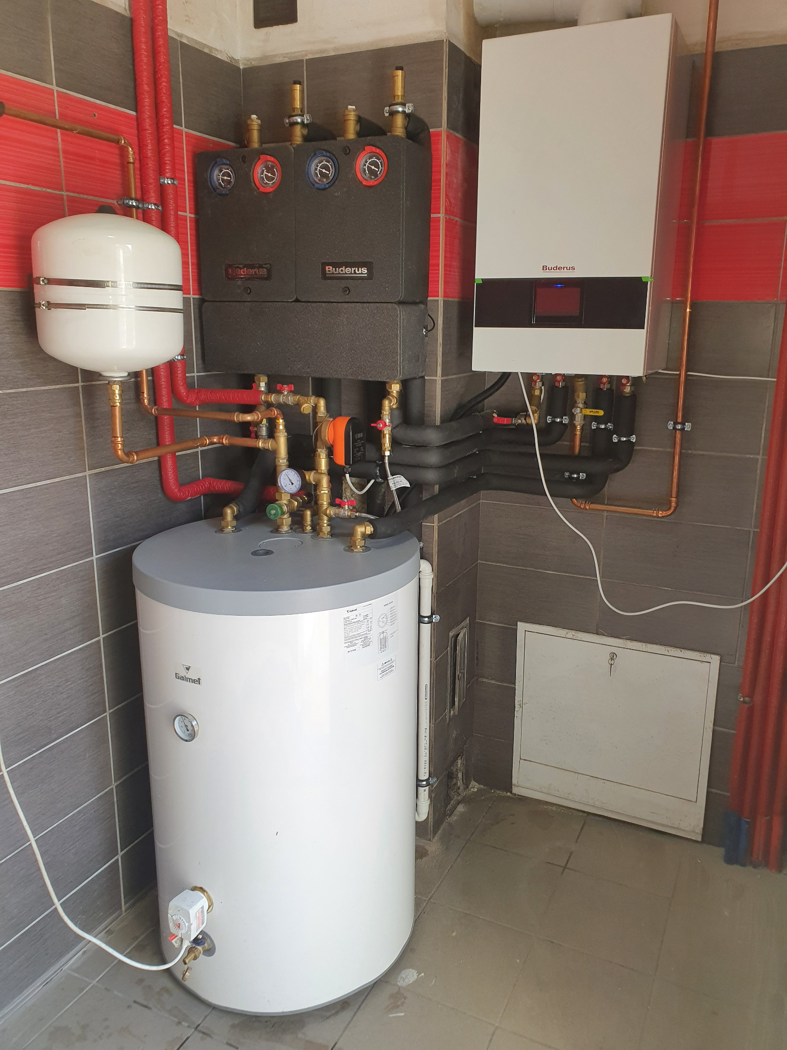 Kompletna instalacja ogrzewania centralnego z kotłem gazowym Buderus, zasobnikiem Galmet i osprzętem, widok na szare płytki i czerwone rury.