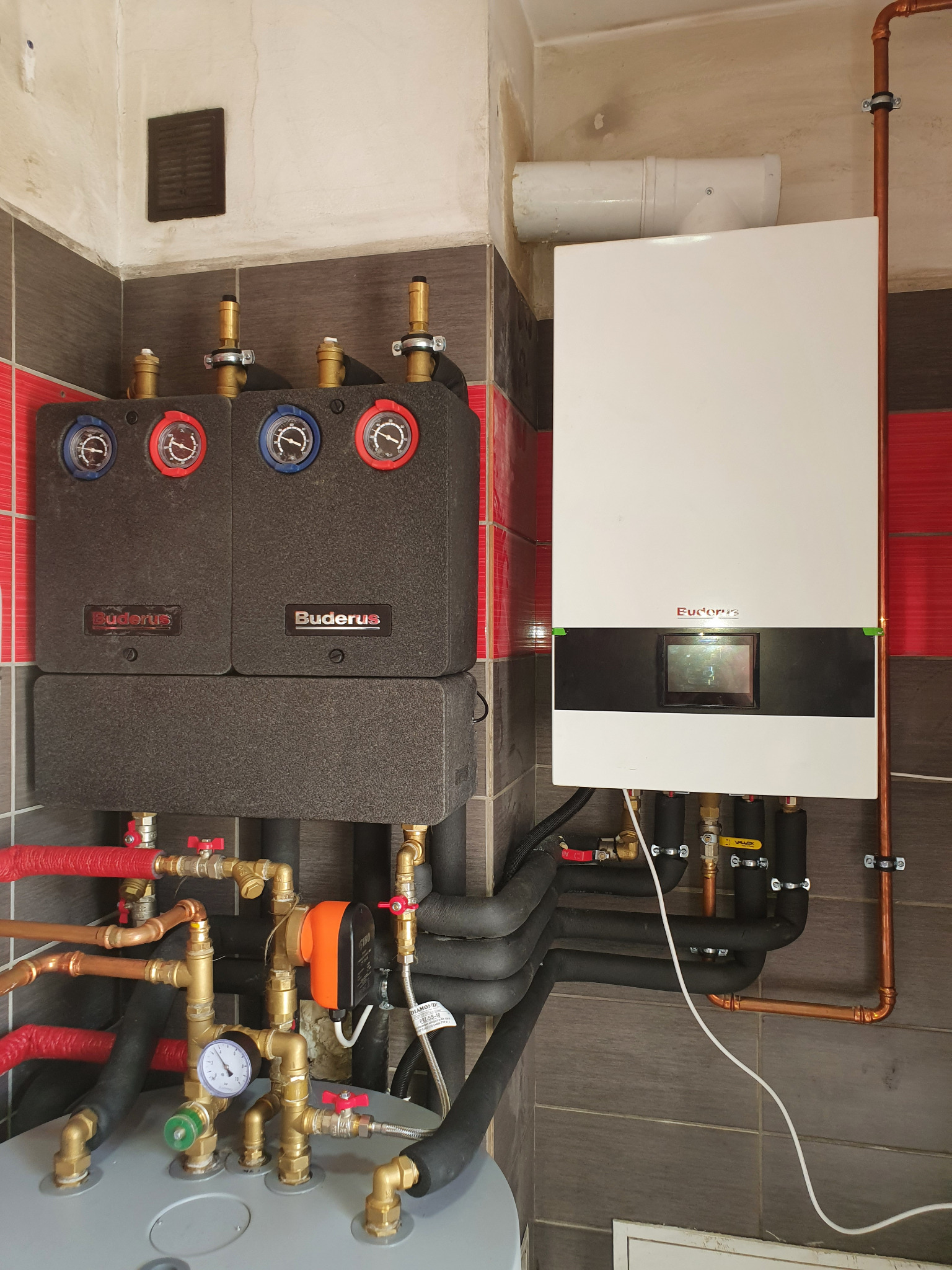 Nowoczesny system grzewczy Buderus z kotłem gazowym Futura, widoczne liczne rury miedziane i zawory, zainstalowany na tle szaro-czerwonej ściany z płytek.