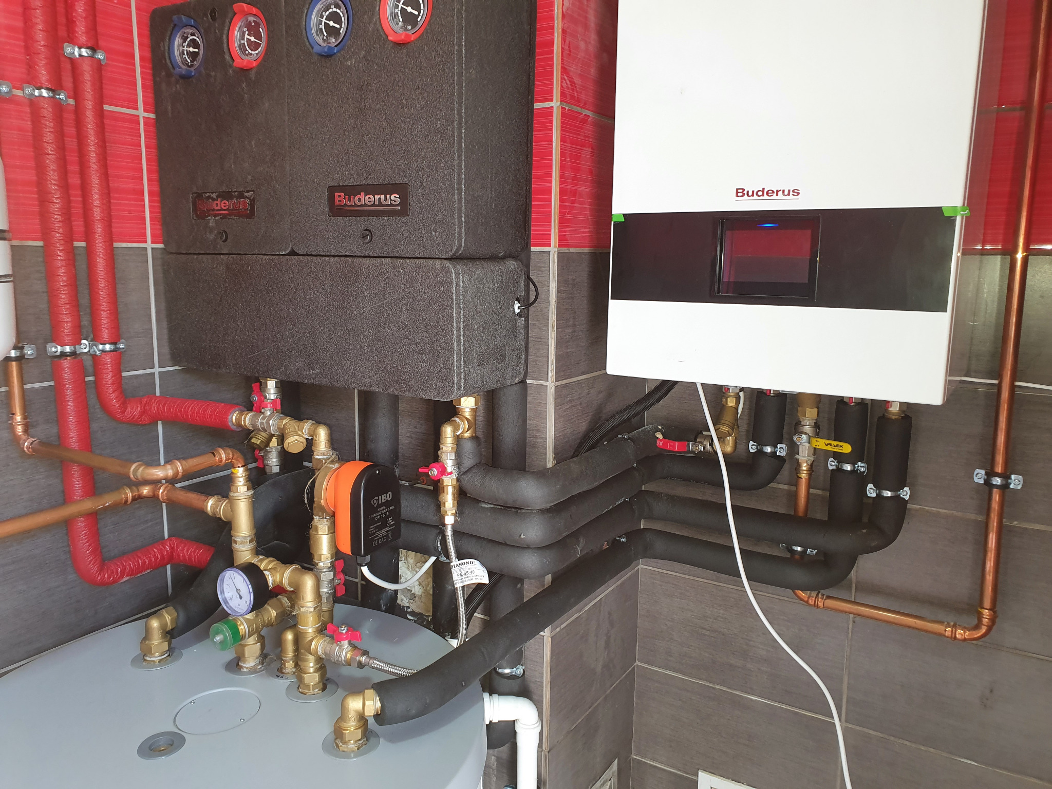 Zestaw urządzeń grzewczych Buderus z widocznymi rurami miedzianymi, czarnymi izolowanymi przewodami, pompą obiegową IBO i manometrem, zamontowany na tle płytek imitujących drewno.