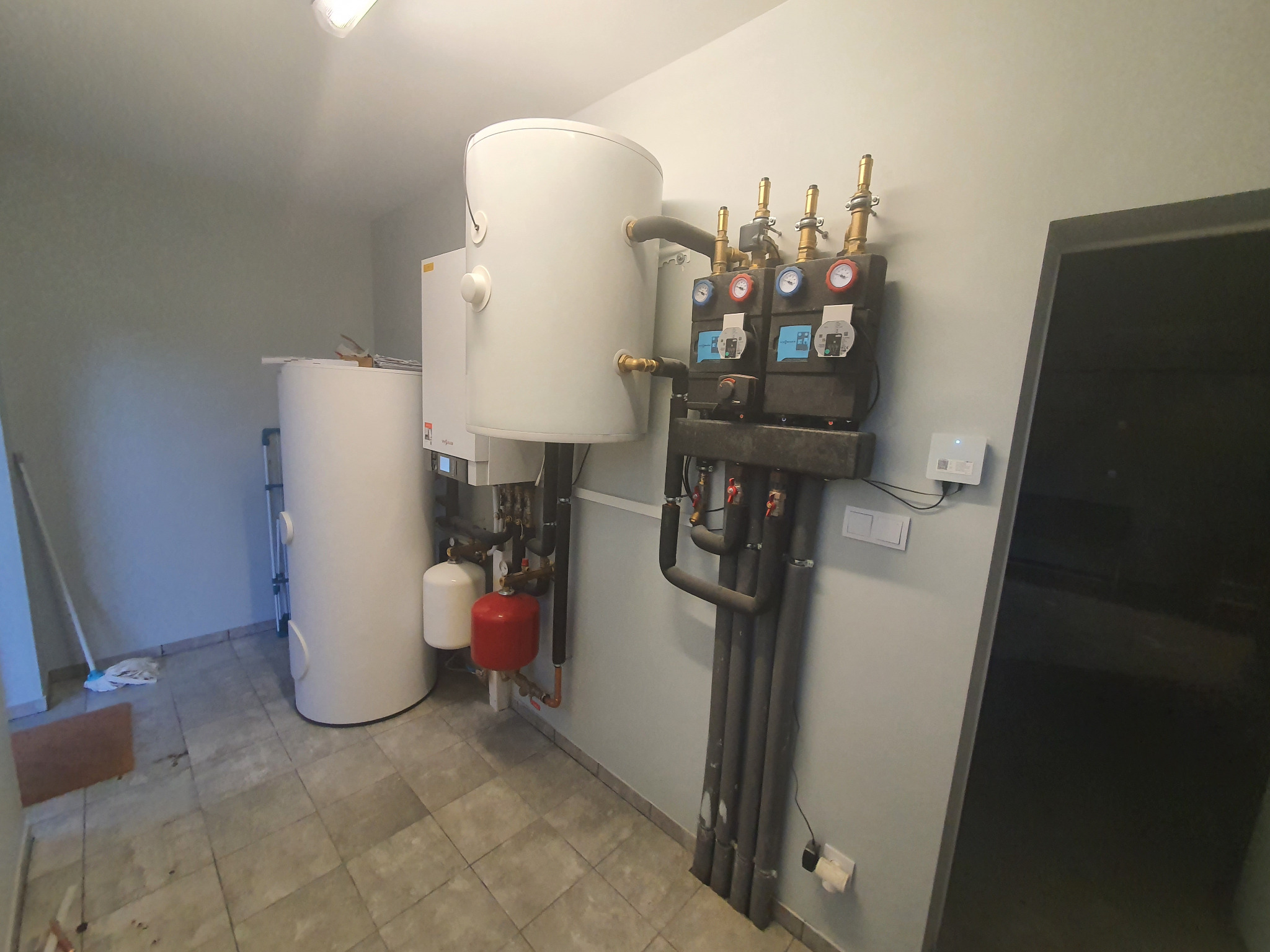 Biały zasobnik ciepłej wody użytkowej z osprzętem i czarną armaturą, zainstalowany w kotłowni z szarą podłogą i ścianami.