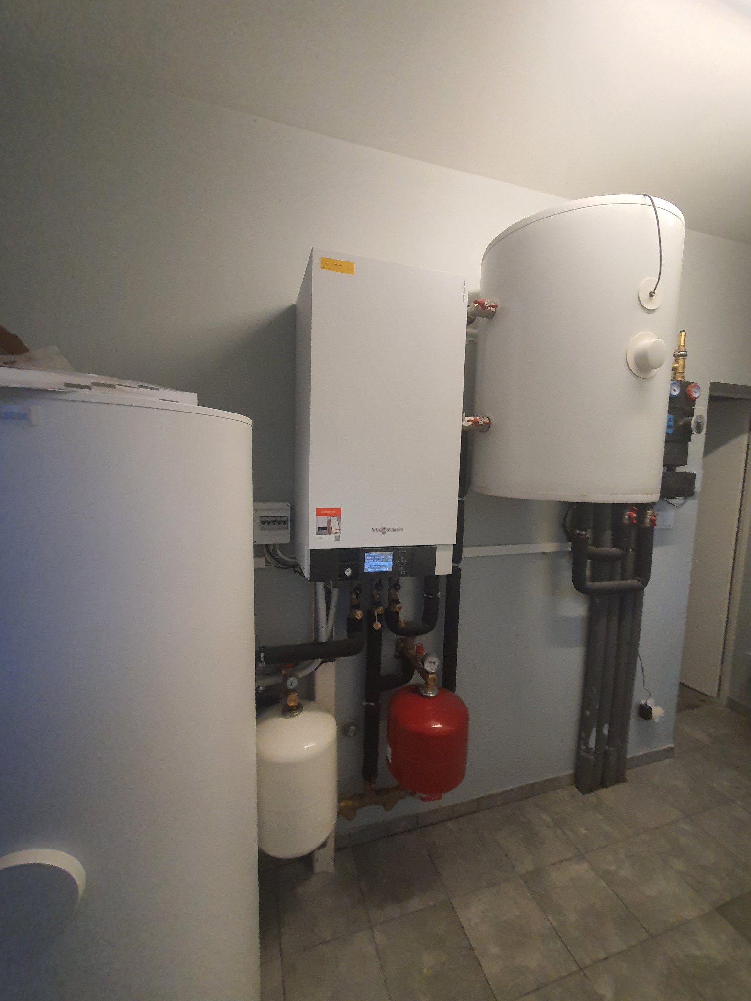 Zestaw instalacji grzewczej z kotłem, zasobnikiem ciepłej wody i naczyniami przeponowymi w pomieszczeniu technicznym z szarą podłogą.