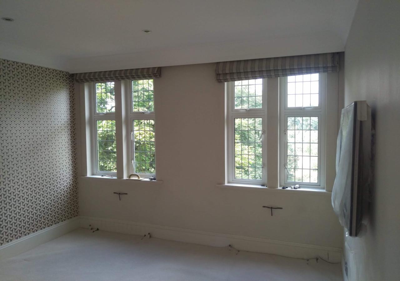 Pokój po remoncie z dwoma oknami z roletami rzymskimi w paski, tapetą w geometryczne wzory na jednej ścianie i wystającymi przewodami elektrycznymi ze ścian.