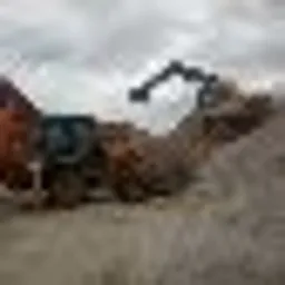 Pomarańczowa koparka nabiera ziemię z hałdy w pochmurny dzień. Widoczne ślady opon na gruncie.