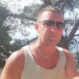 Mężczyzna w okularach przeciwsłonecznych i białym podkoszulku na tle drzewa i morza. Widoczny łańcuszek z krzyżykiem.