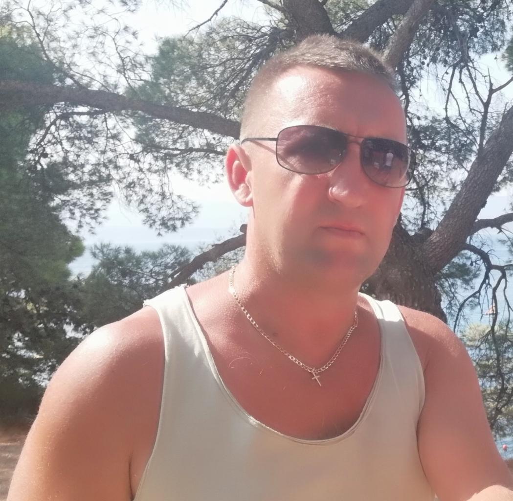 Mężczyzna w okularach przeciwsłonecznych i białym podkoszulku na tle drzewa i morza. Widoczny łańcuszek z krzyżykiem.