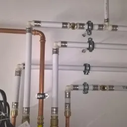 Fragment instalacji wodnej i grzewczej z rur miedzianych i plastikowych, zamocowanych do ściany za pomocą metalowych uchwytów. Widoczne złącza, kolanka, przewody elektryczne i fragment etykiety...