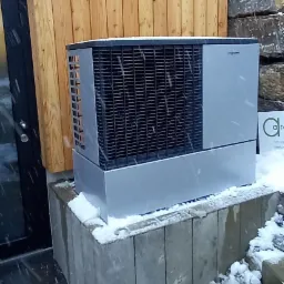 Zewnętrzna pompa ciepła Viessmann zamontowana na betonowym cokole, lekko ośnieżona podczas opadów śniegu, na tle drewnianej elewacji budynku.