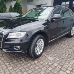 Błyszczący, czarny Audi Q5, zaparkowany na kostce brukowej przed budynkiem z szarą elewacją. Widoczny fragment tablicy rejestracyjnej i srebrne felgi. W tle zaparkowany ciemny samochód.