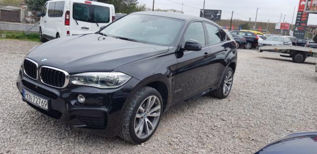 Czarny samochód typu SUV marki BMW, model X6, zaparkowany na żwirowym parkingu z widocznym numerem rejestracyjnym, białym vanem w tle i przyczepą samochodową po prawej stronie.