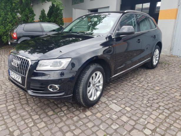 Błyszczący, czarny Audi Q5, zaparkowany na kostce brukowej przed budynkiem z szarą elewacją. Widoczny fragment tablicy rejestracyjnej i srebrne felgi. W tle zaparkowany ciemny samochód.