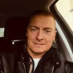 Mężczyzna w skórzanej kurtce siedzi w samochodzie, patrzy w obiektyw z delikatnym uśmiechem.