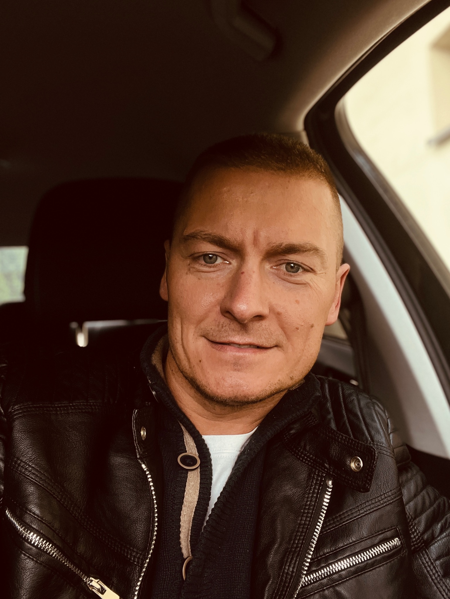 Mężczyzna w skórzanej kurtce siedzi w samochodzie, patrzy w obiektyw z delikatnym uśmiechem.