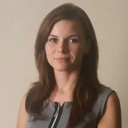 Portret uśmiechniętej kobiety z brązowymi włosami w szarej sukience z zamkiem błyskawicznym na tle kremowej ściany.