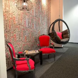 Aranżacja biura: Dwa czerwone fotele w stylu retro, czarny stolik z gazetą, wiszące krzesło-huśtawka i ściana z motywem napisu NYC