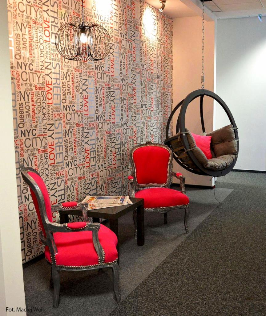 Aranżacja biura: Dwa czerwone fotele w stylu retro, czarny stolik z gazetą, wiszące krzesło-huśtawka i ściana z motywem napisu NYC