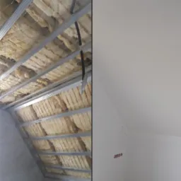 Poddasze w trakcie adaptacji: widok na izolację z wełny mineralnej między metalowymi profilami sufitu, z prawej fragment ściany po szpachlowaniu i malowaniu na biało.