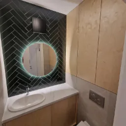 Nowoczesna łazienka z umywalką nablatową, szafką z jasnego drewna, okrągłym lustrem z podświetleniem LED na ścianie wyłożonej ciemnozieloną płytką w jodełkę oraz zabudową z jasnego drewna.