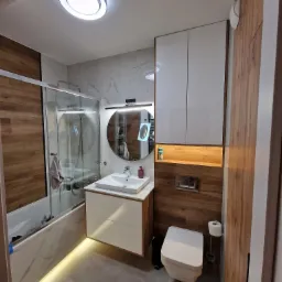 Nowoczesna łazienka z wanną za szklaną ścianką, umywalką z szafką, okrągłym lustrem z podświetleniem LED, podwieszaną toaletą i drewnopodobnymi panelami na ścianach, oświetleniem LED pod wanną i...