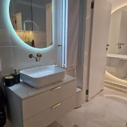 Nowoczesna łazienka z białą umywalką nablatową, armaturą w kolorze złotym, szafką z marmurowym blatem i podświetlanym lustrem w kształcie koła, z widocznym fragmentem grzejnika.