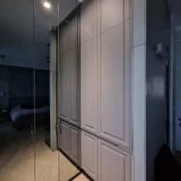 Szara szafa wnękowa z lustrzanymi drzwiami, odbijająca parkiet w jodełkę i fragment sypialni z ciemną tapetą 3D na ścianie.