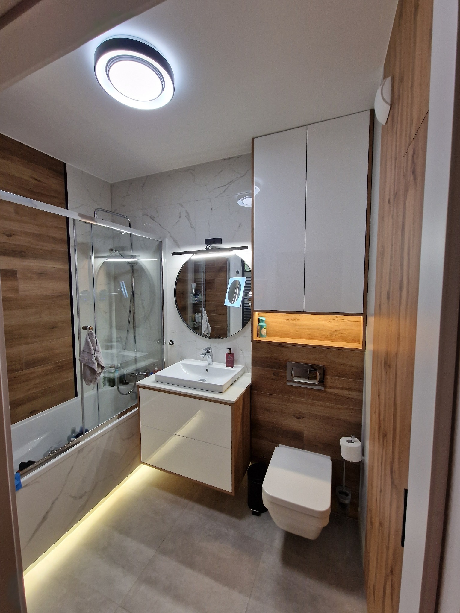 Nowoczesna łazienka z wanną za szklaną ścianką, umywalką z szafką, okrągłym lustrem z podświetleniem LED, podwieszaną toaletą i drewnopodobnymi panelami na ścianach, oświetleniem LED pod wanną i...