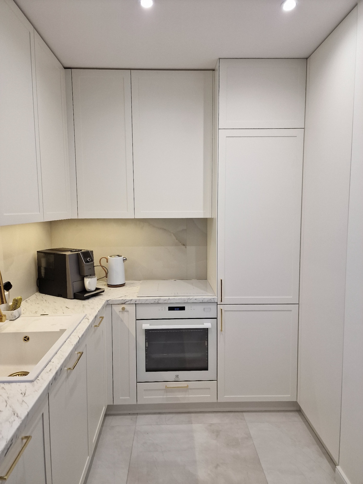 Jasna kuchnia z białymi frontami, marmurowym blatem i złotymi uchwytami, wyposażona w ekspres do kawy i czajnik.