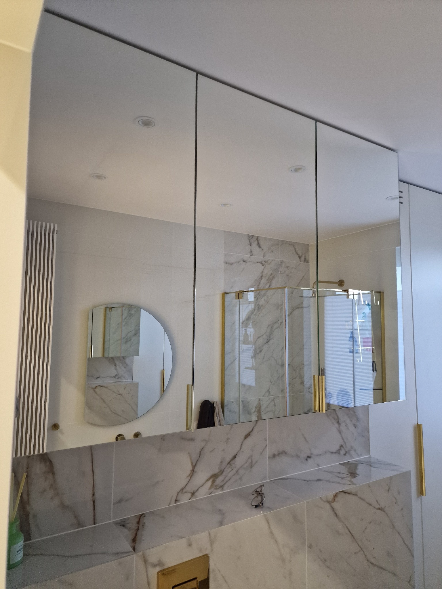 Nowoczesna łazienka z trzema lustrzanymi szafkami wiszącymi, marmurowym blatem i okrągłym lustrem na ścianie, odbijającym kabinę prysznicową z złotymi detalami.