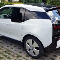 Biały samochód elektryczny BMW i3 z przyciemnionymi szybami, zaparkowany na betonowej kostce z trawą, widok z boku i tyłu, z widocznym logo BMW na kole i tylnej klapie.