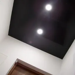 Czarne, błyszczące wykończenie sufitu z dwoma wbudowanymi punktami świetlnymi, widok od dołu, z fragmentem drewnianych drzwi z metalową wstawką w tle.