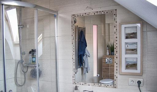 Ukończona łazienka na poddaszu z kabiną prysznicową ze szklanymi drzwiami, lustrem w mozaikowej ramie, wiszącą toaletą i dekoracyjną ramką z trzema obrazkami plażowymi.