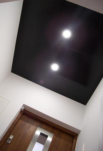 Czarne, błyszczące wykończenie sufitu z dwoma wbudowanymi punktami świetlnymi, widok od dołu, z fragmentem drewnianych drzwi z metalową wstawką w tle.