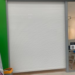 PHU SUN-TEL Cezary Dobrzyński - Zasunięta biała roleta antywłamaniowa w lokalu handlowym, widoczny fragment reklamy i kosz na śmieci obok.