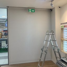 PHU SUN-TEL Cezary Dobrzyński - Zamknięta, biała roleta antywłamaniowa w sklepie, widoczna aluminiowa drabina oparta o roletę, w tle regał z okularami i wyjście ewakuacyjne.