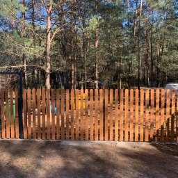 PHU SUN-TEL Cezary Dobrzyński - Nowe drewniane ogrodzenie z furtką, widok z zewnątrz, w tle las iglasty i zaparkowany biały samochód dostawczy.