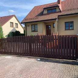 Brązowa, przesuwna brama wjazdowa z pionowych sztachet, zamontowana na posesji z dwoma domami o beżowych elewacjach i brązowych dachach, widok z zewnątrz.