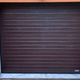 PHU SUN-TEL Cezary Dobrzyński - Zamknięta brama garażowa segmentowa w kolorze ciemnego drewna, z poziomymi przetłoczeniami, zamontowana w pomarańczowym budynku. Widoczny fragment muru z cegły.