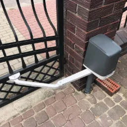 Czarna, metalowa brama wjazdowa z zamontowaną automatyką (szary napęd z białym ramieniem) przy słupku z czerwonej cegły, na tle kostki brukowej.